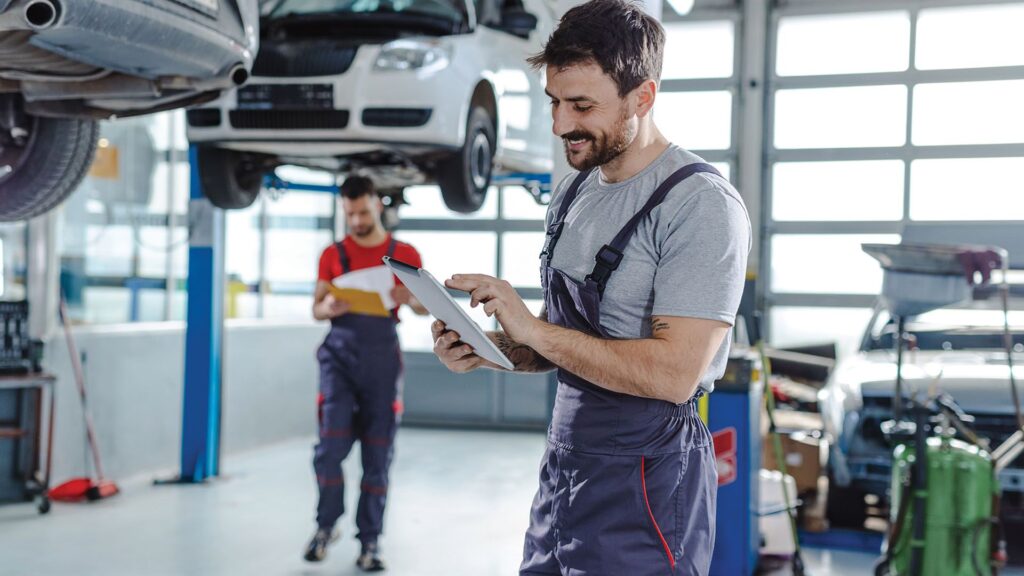 A mechanic is using a tablet in a garage while another mechanic looks at a clipboard